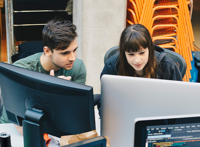 Two business cybersecurity professional look over their work on two computer monitors.