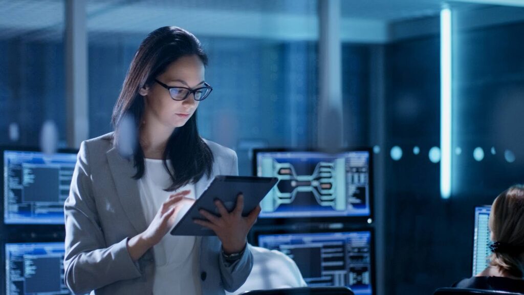 A young woman in a system control center uses a tablet; multiple screens, some occupied by coworkers, are visible in the background.