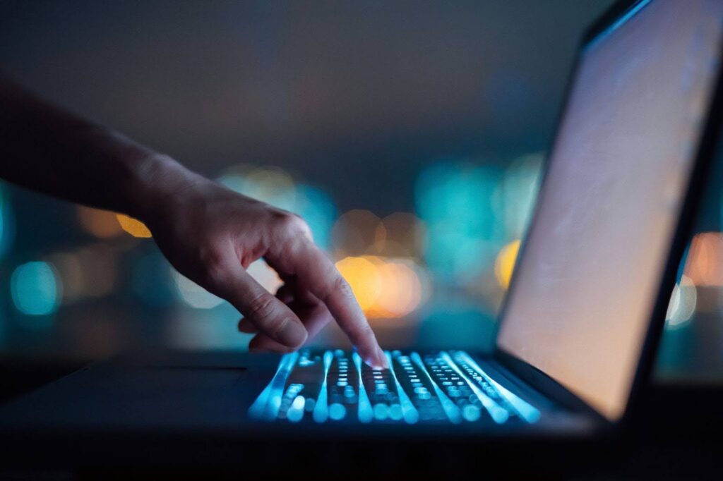 Close up of a woman's hand typing on a computer keyboard in the dark against a colorful background bokeh.