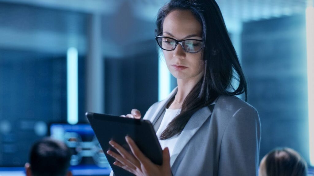 A bespectacled woman in business attire uses a tablet computer; in the background, coworkers working at computer stations are visible.