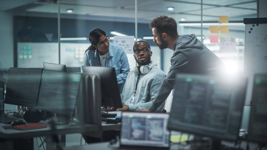 Three professionals in a business office computing center converse at a workstation where one is seated.