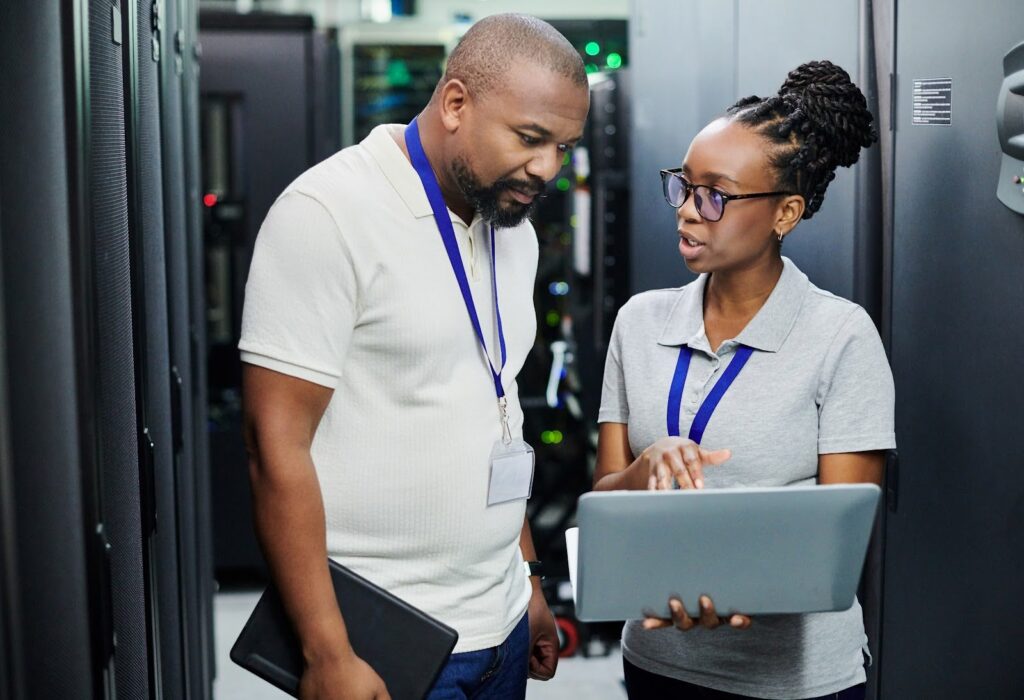 Two computing professionals, one holding a laptop computer, confer in a server room.