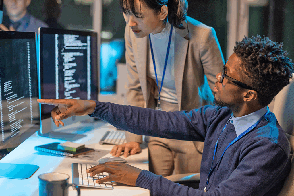 A business cybersecurity consultant is sitting, showing code on a computer screen to a colleague observing attentively while standing next to him.